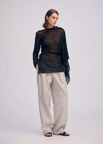 A woman stands against a plain light background, hands in pockets, confidently wearing the ISSEY MIYAKE CONCEALED KNIT sheer sparkly long-sleeved top over a black bra, paired with loose beige trousers and black open-toe sandals.