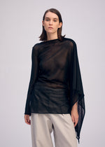 A woman with straight brown hair wears the ISSEY MIYAKE CONCEALED KNIT, a sheer sparkly nylon long-sleeved top with an asymmetrical design, and loose beige trousers. She stands against a plain background, looking at the camera with a neutral expression.