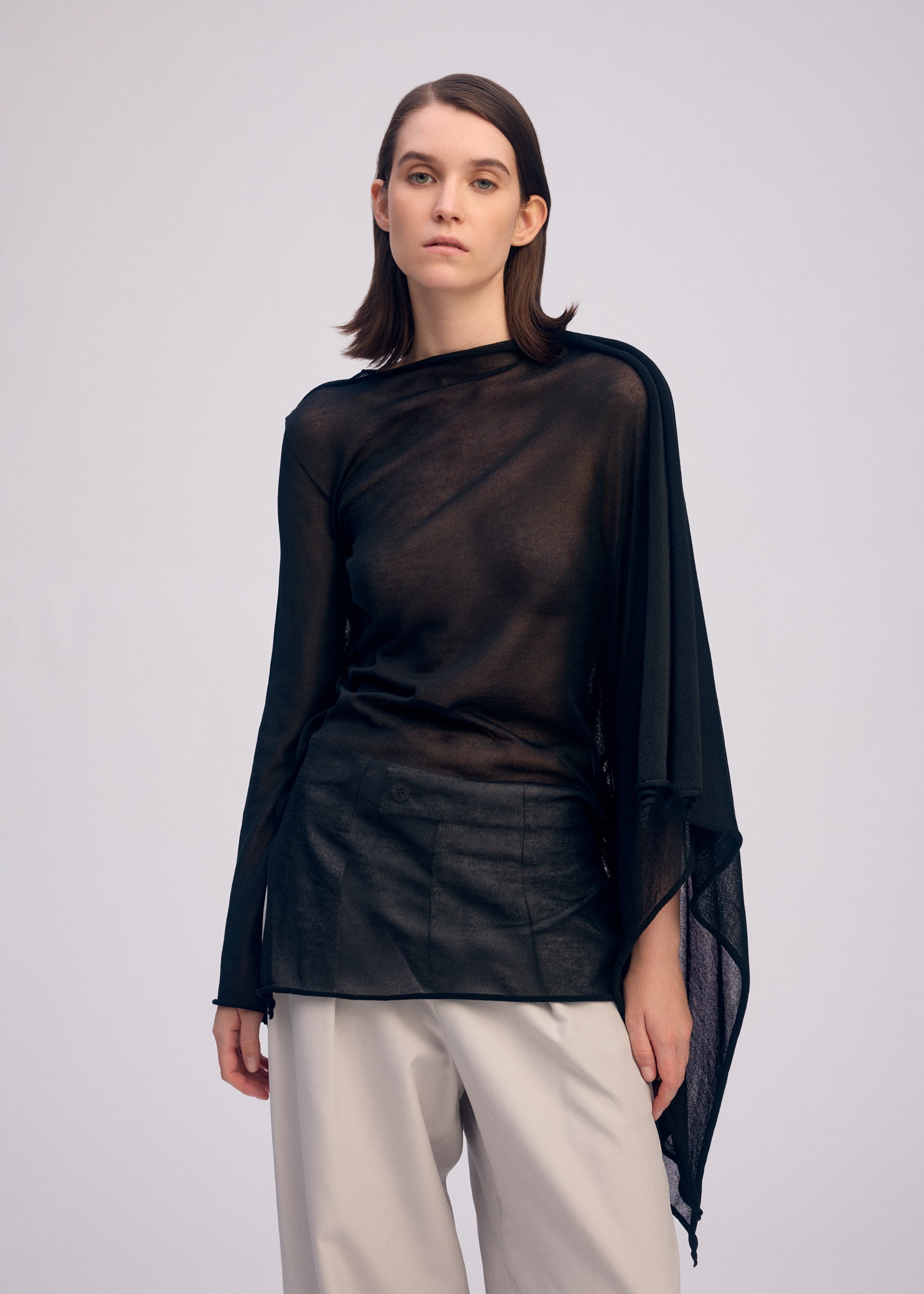 Zoomed view A woman with straight brown hair wears the ISSEY MIYAKE CONCEALED KNIT, a sheer sparkly nylon long-sleeved top with an asymmetrical design, and loose beige trousers. She stands against a plain background, looking at the camera with a neutral expression.