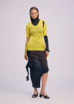 A woman models ISSEY MIYAKE’s ADVENTITIOUS：SLEEVES—a yellow knit top over a black turtleneck and sheer abstract midi skirt—styled with black heeled sandals against a plain backdrop.