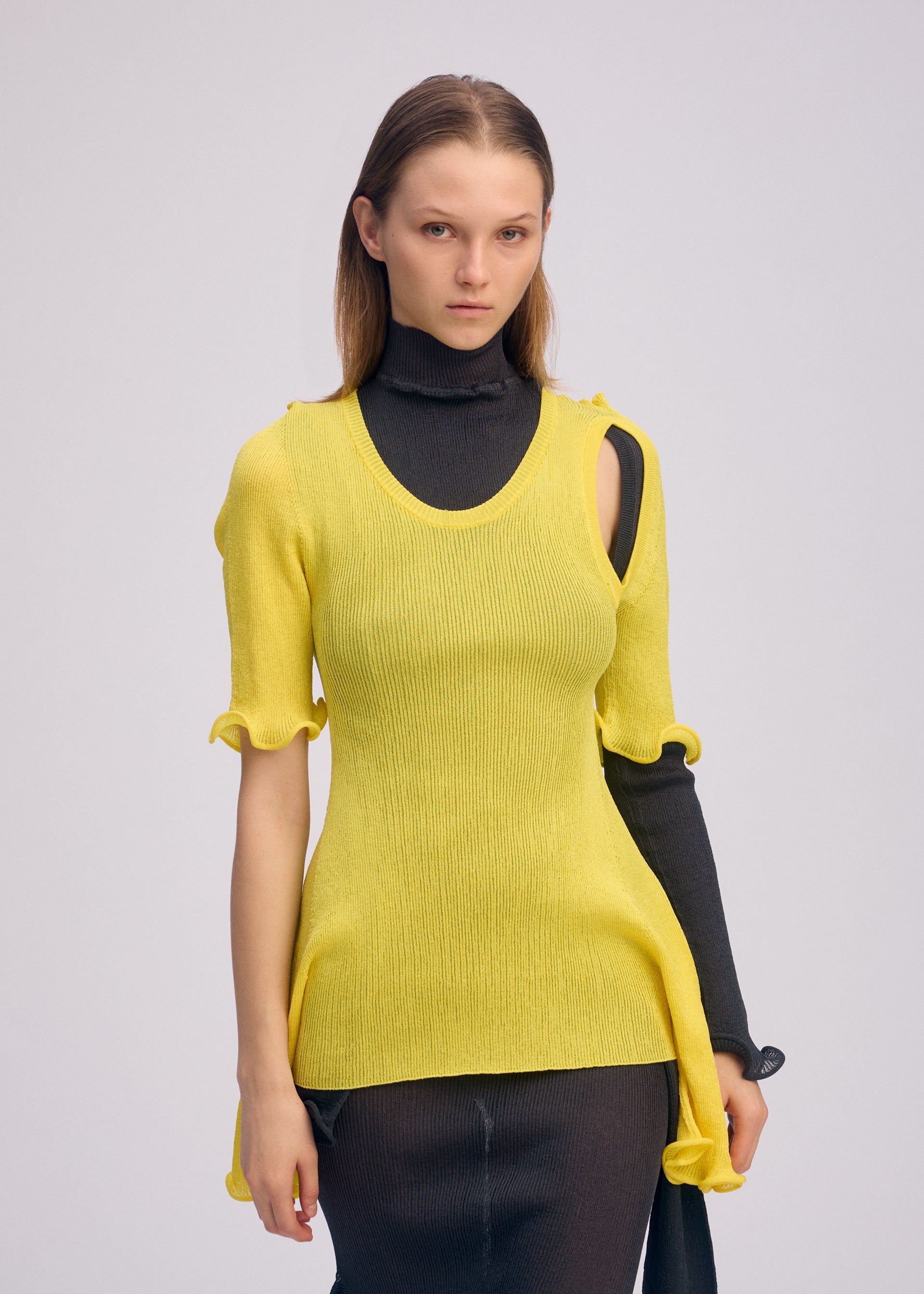 Zoomed view A woman with straight brown hair models ISSEY MIYAKE’s ADVENTITIOUS: SLEEVES—a fitted yellow knit top with ruffles layered over a long black turtleneck dress, exposing one sleeve for an asymmetrical, versatile look against a plain background.