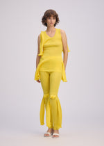 A person with short curly hair wears ISSEY MIYAKE's ADVENTITIOUS: SLEEVES—a sleeveless yellow knit top with asymmetrical draping, paired with matching flared yellow pants and white sandals, standing against a plain light background.