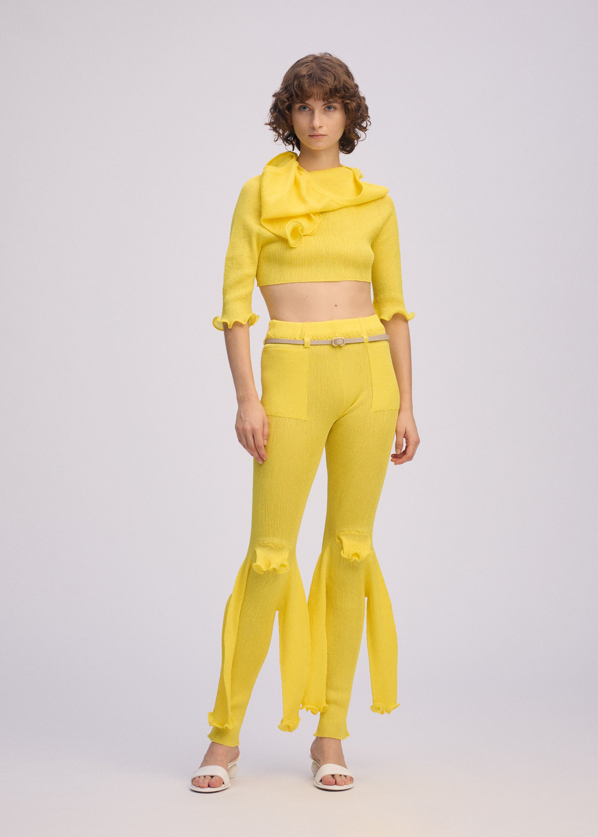 Zoomed view A woman models the ISSEY MIYAKE ADVENTITIOUS：SLEEVES—a bright yellow knit top with ruffled sleeves and matching flared pants—styled with her short curly hair and white sandals against a plain backdrop.
