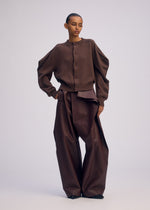 A person stands against a plain background wearing the ISSEY MIYAKE AKIMBO KNIT, an oversized cardigan with puffy-shoulder sleeves, paired with high-waisted, wide-leg brown pants featuring an asymmetrical flap detail.