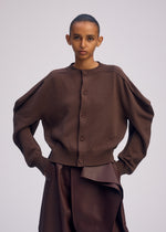 A person with slicked-back hair models ISSEY MIYAKE’s AKIMBO KNIT, a brown organic cotton cardigan with wide sleeves, paired with a high-waisted brown skirt with an asymmetrical front detail, set against a plain light background.