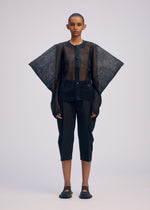 A person with short hair and a neutral expression wears the CIALDA sheer black button-up top by ISSEY MIYAKE, featuring wide, wing-like sleeves. They pair it with black cropped pants and black shoes against a plain background.