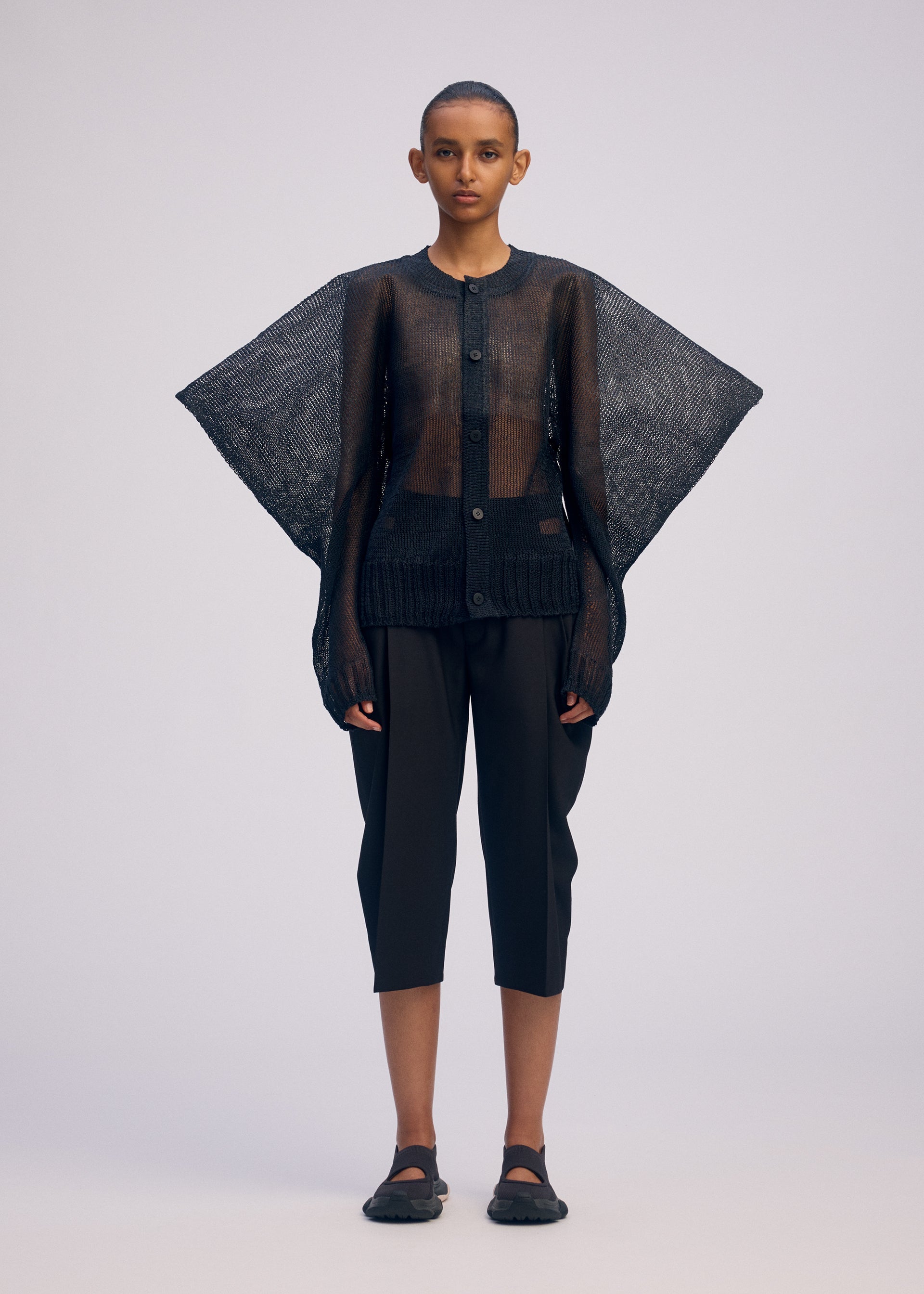 Zoomed view A person with short hair and a neutral expression wears the CIALDA sheer black button-up top by ISSEY MIYAKE, featuring wide, wing-like sleeves. They pair it with black cropped pants and black shoes against a plain background.