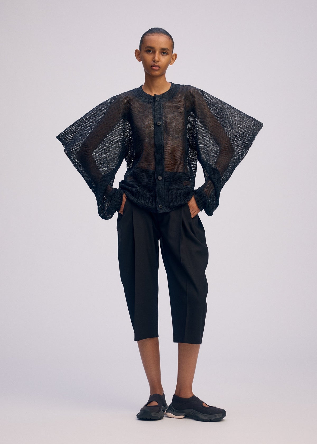 A person stands facing forward wearing the ISSEY MIYAKE CIALDA sheer black button-up top with wide sleeves, paired with black knee-length shorts and slip-on shoes. The plain background highlights the outfit’s structure and texture. view 6 of 9