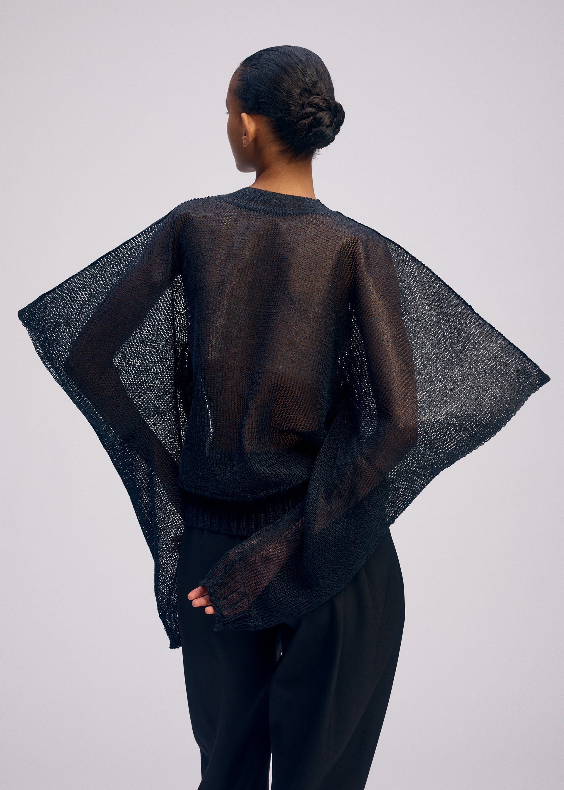 Zoomed view Facing away, a person with hair in a low bun wears the ISSEY MIYAKE CIALDA sheer mesh-textured black top with wide sleeves and black pants. One hand is behind their back against a plain, light background.