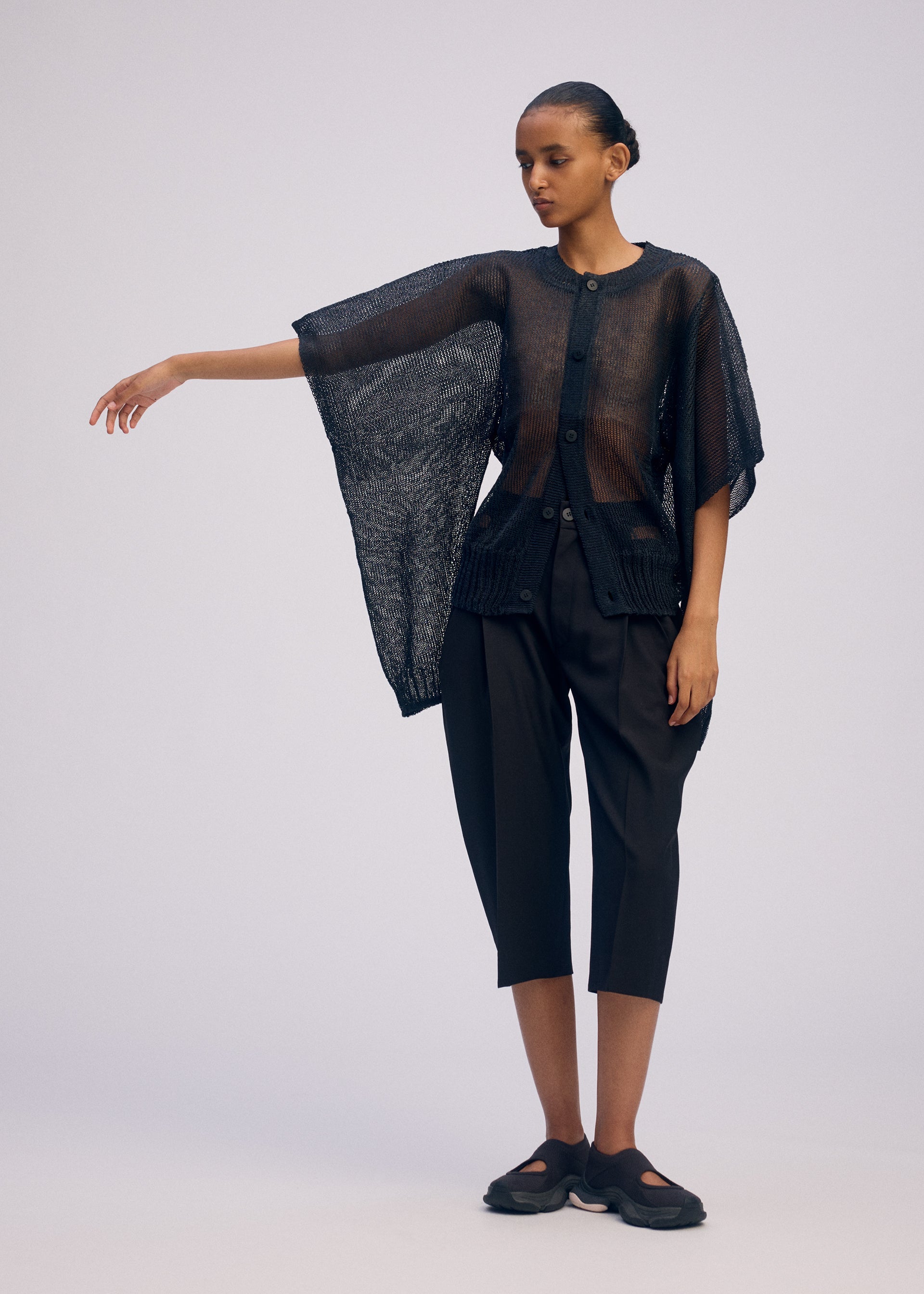 Zoomed view Against a plain background, a person with short, slicked-back hair and a neutral expression wears ISSEY MIYAKE's CIALDA: a sheer black mesh-like top with wide sleeves, paired with black cropped pants and black shoes.