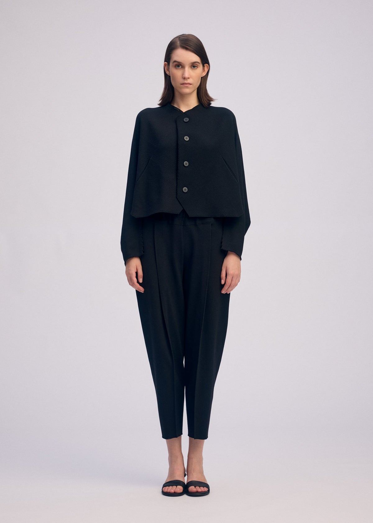 A woman stands facing forward, wearing the CAMPAGNE black button-up cape-style top from ISSEY MIYAKE, crafted from stretchy material, paired with black tapered pants and black sandals against a plain light background. view 3 of 5