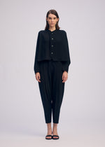 A woman stands facing forward, wearing the CAMPAGNE black button-up cape-style top from ISSEY MIYAKE, crafted from stretchy material, paired with black tapered pants and black sandals against a plain light background.