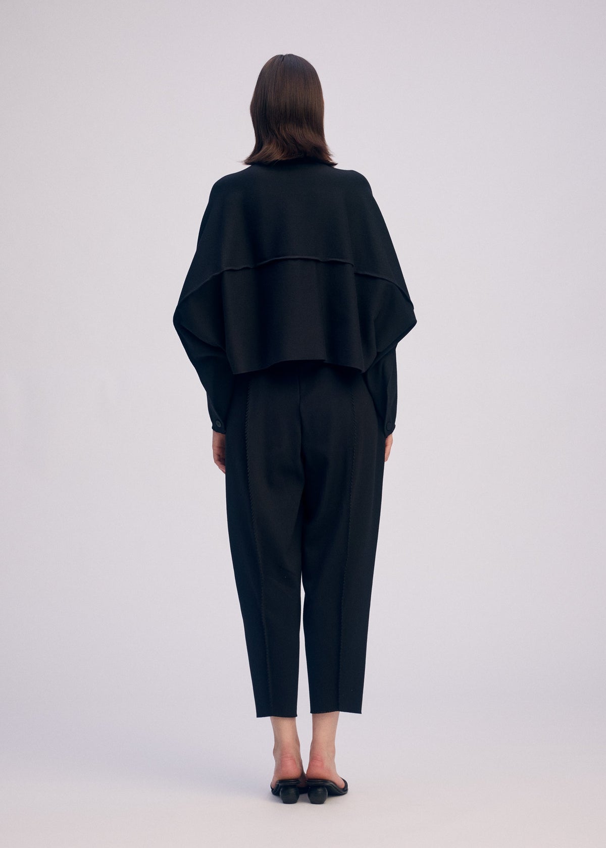 Zoomed view A person with straight brown hair stands barefoot, facing away from the camera against a plain light background, wearing loose black CAMPAGNE pants made from stretchy material by ISSEY MIYAKE, paired with a long-sleeve top and a layered black cape.