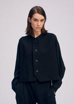 A woman with straight brown hair models the ISSEY MIYAKE CAMPAGNE ensemble—a loose-fitting, asymmetrical black jacket made from stretchy fabric with large buttons, paired with matching black pants—standing against a light gray background.