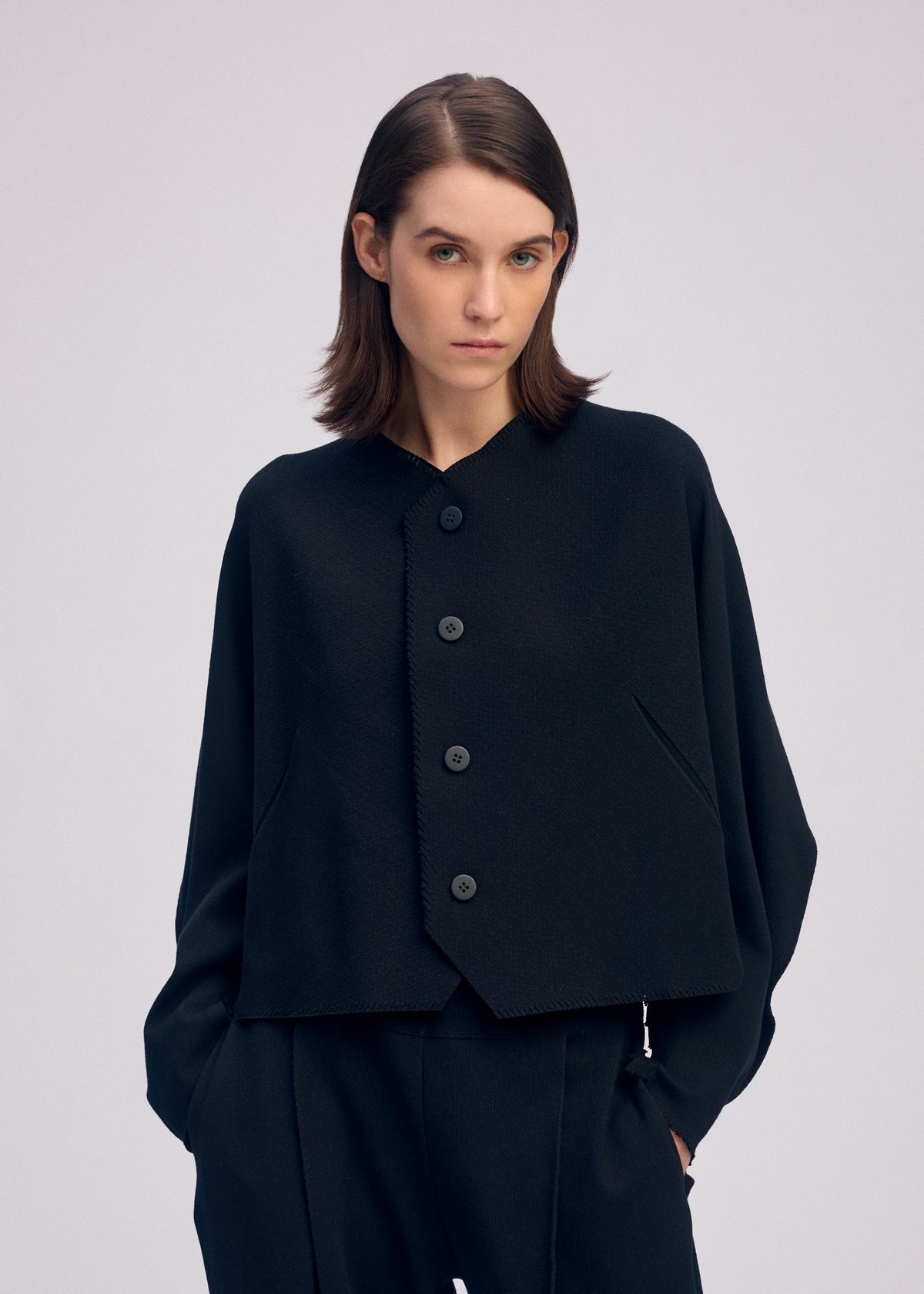Zoomed view A woman with straight brown hair models the ISSEY MIYAKE CAMPAGNE ensemble—a loose-fitting, asymmetrical black jacket made from stretchy fabric with large buttons, paired with matching black pants—standing against a light gray background.