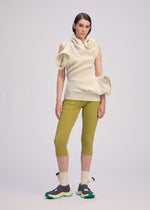 A woman stands against a light backdrop, wearing the sculptural off-white LIFE FORM knit tunic by ISSEY MIYAKE, yellow-green cropped pants, beige socks, and black sandals with green details. She faces the camera.
