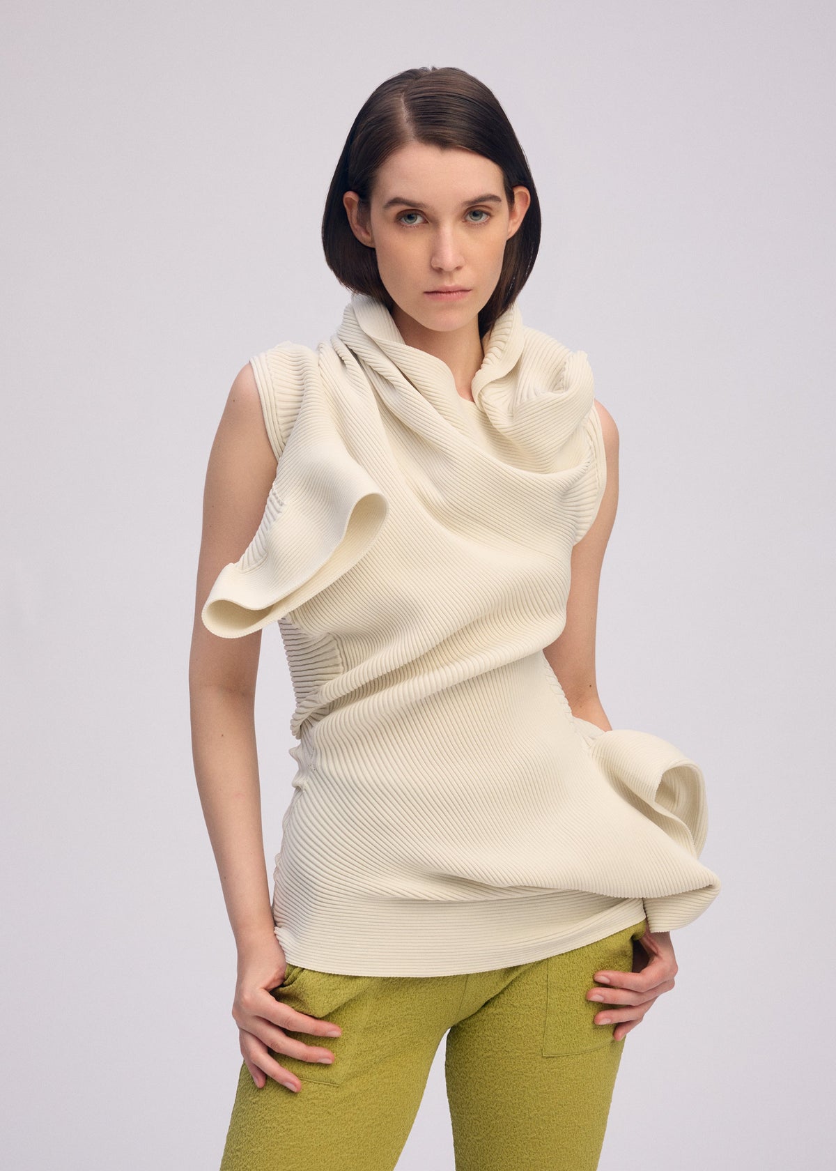 A woman with a neutral expression wears the ISSEY MIYAKE LIFE FORM ivory knit tunic with textured, wavy folds and olive-green pants, standing against a plain light gray background. view 6 of 7