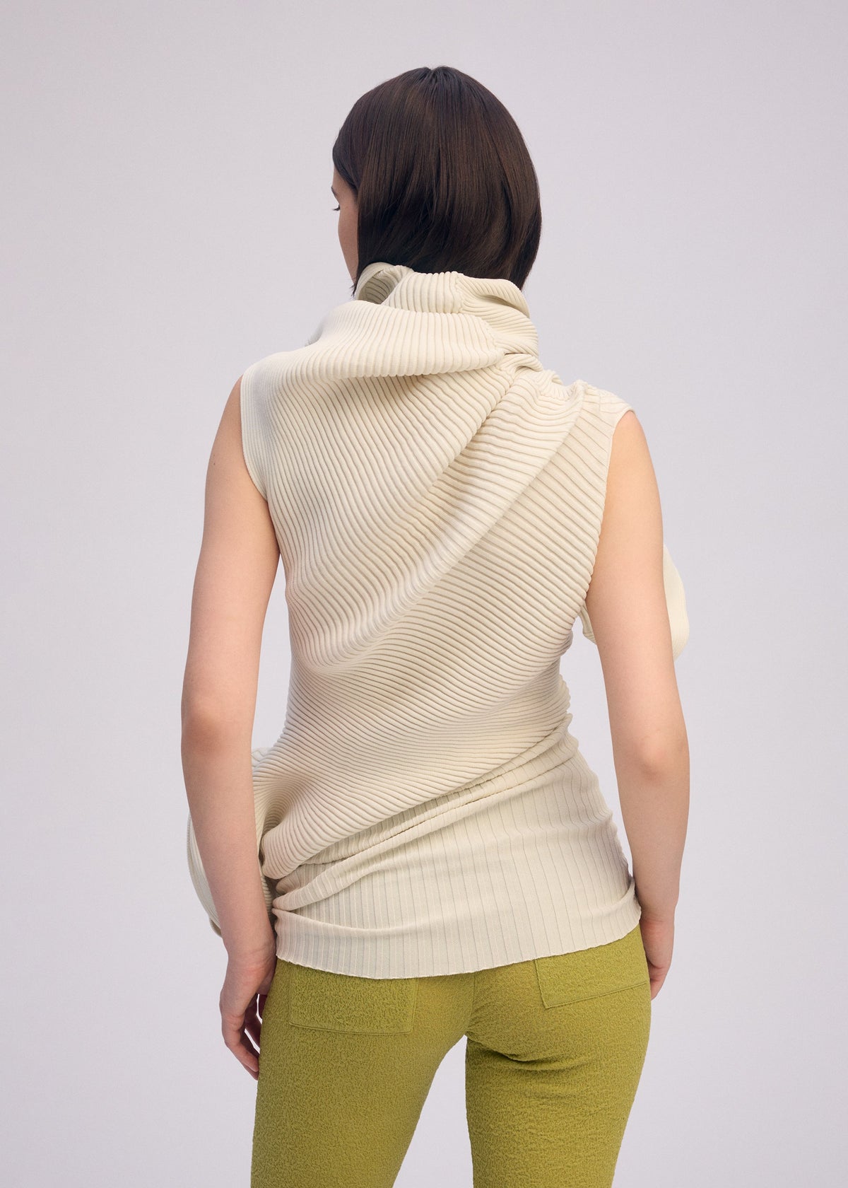 A person with straight, dark brown hair faces away, wearing ISSEY MIYAKE’s LIFE FORM: a sleeveless cream knit tunic with draped collar and light olive green textured pants, set against a plain light background. view 7 of 7