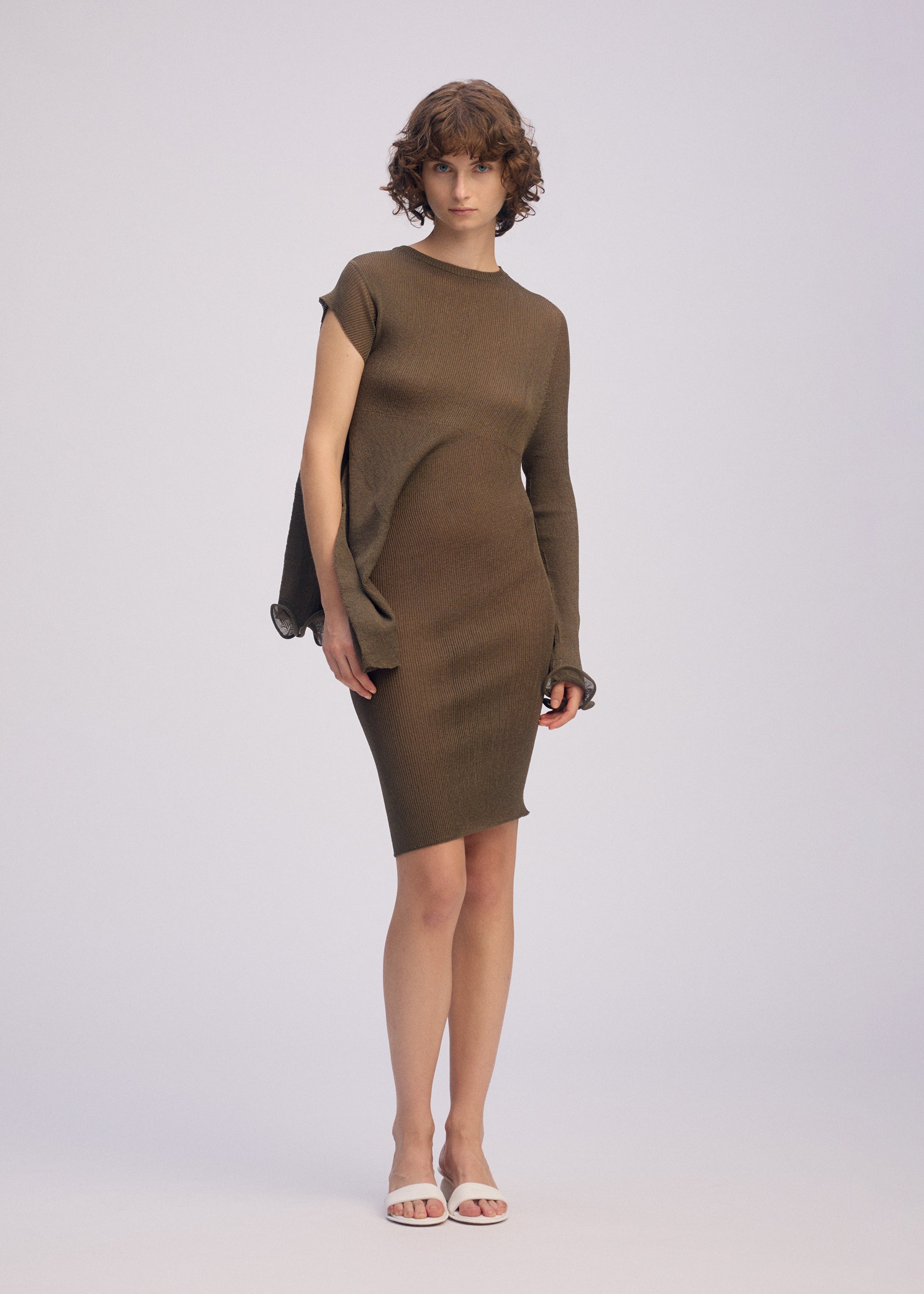 Zoomed view A person with short curly hair wears the ISSEY MIYAKE ADVENTITIOUS：SLEEVES olive-green asymmetrical knit tunic, featuring one long sleeve and a sleeveless side, styled with white open-toe sandals against a plain light background.