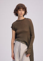 A young person with short, curly brown hair wears the ISSEY MIYAKE ADVENTITIOUS: SLEEVES asymmetric olive-green tunic—crafted with a non-sewn knitting technique—paired with high-waisted, pleated beige trousers against a plain background.