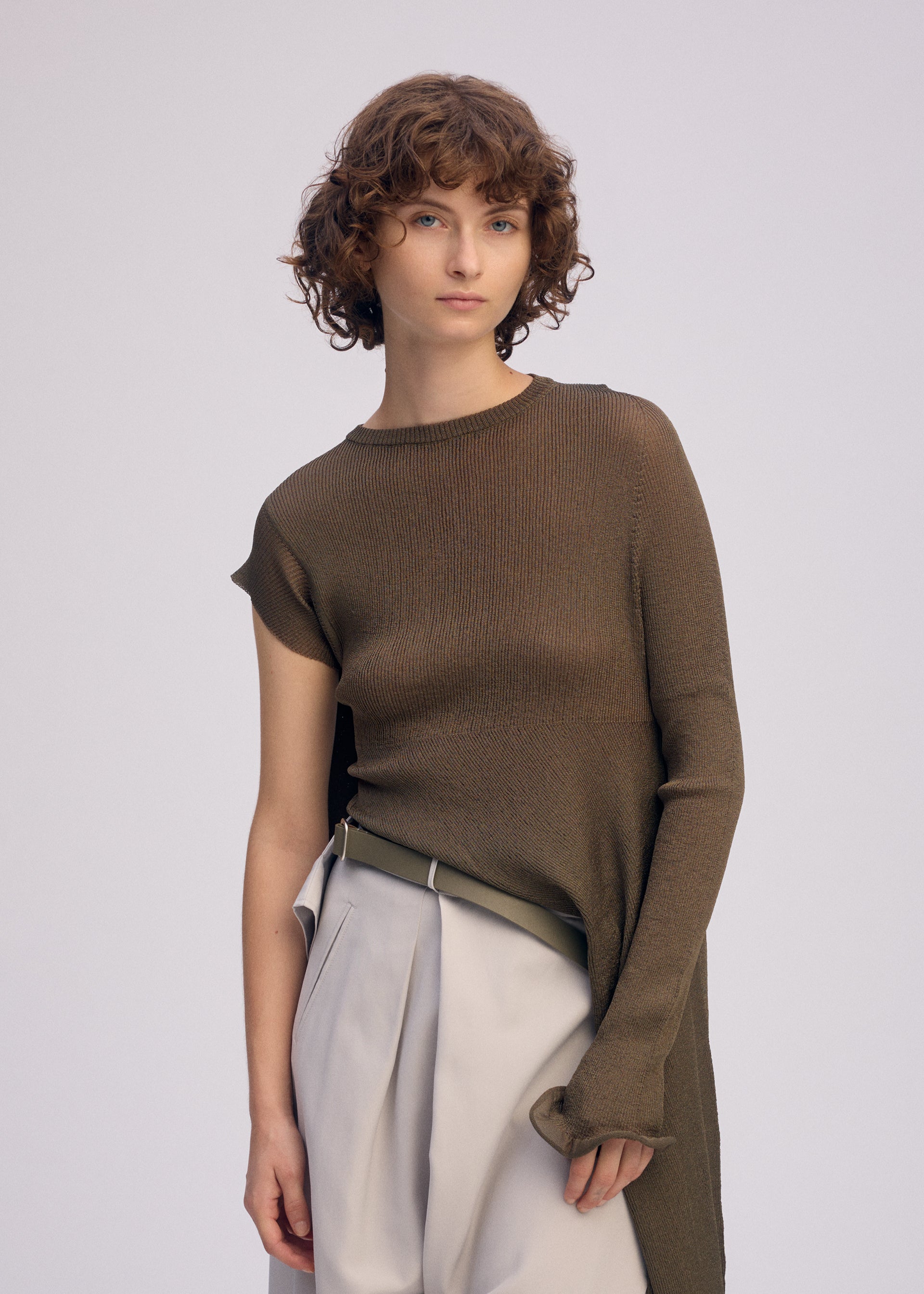 Zoomed view A young person with short, curly brown hair wears the ISSEY MIYAKE ADVENTITIOUS: SLEEVES asymmetric olive-green tunic—crafted with a non-sewn knitting technique—paired with high-waisted, pleated beige trousers against a plain background.