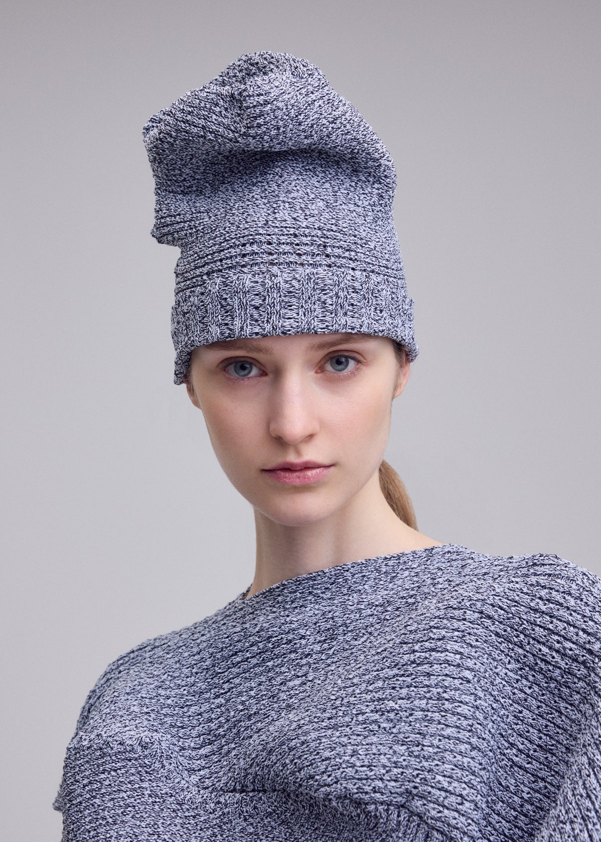A person with fair skin and blue eyes wears the ISSEY MIYAKE FOUND STONE BEANIE and matching top, looking directly at the camera against a plain light gray background. view 4 of 5