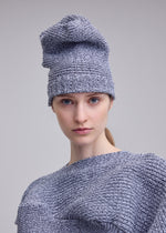 A person with fair skin and blue eyes wears the ISSEY MIYAKE FOUND STONE BEANIE and matching top, looking directly at the camera against a plain light gray background.