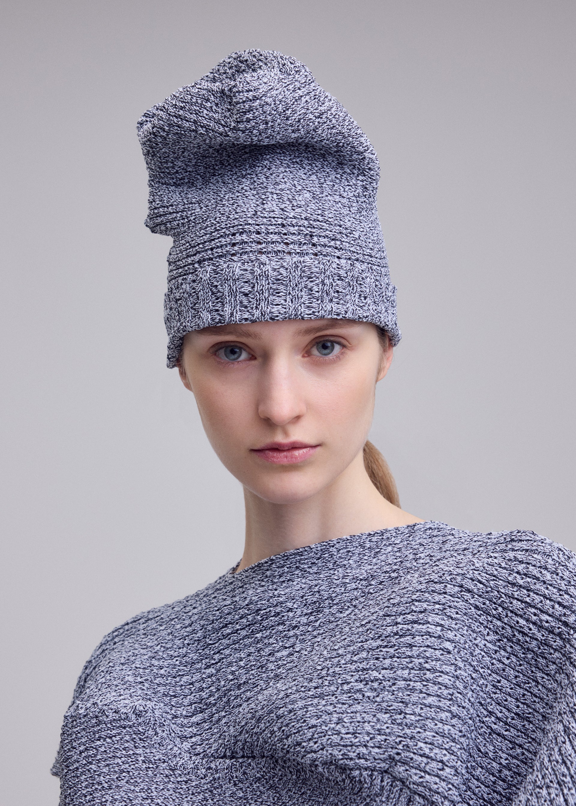 Zoomed view A person with fair skin and blue eyes wears the ISSEY MIYAKE FOUND STONE BEANIE and matching top, looking directly at the camera against a plain light gray background.