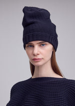 A woman with straight brown hair wears the ISSEY MIYAKE FOUND STONE BEANIE, a three-dimensional, tall, pointed knit hat, paired with a matching top while looking at the camera against a neutral background.