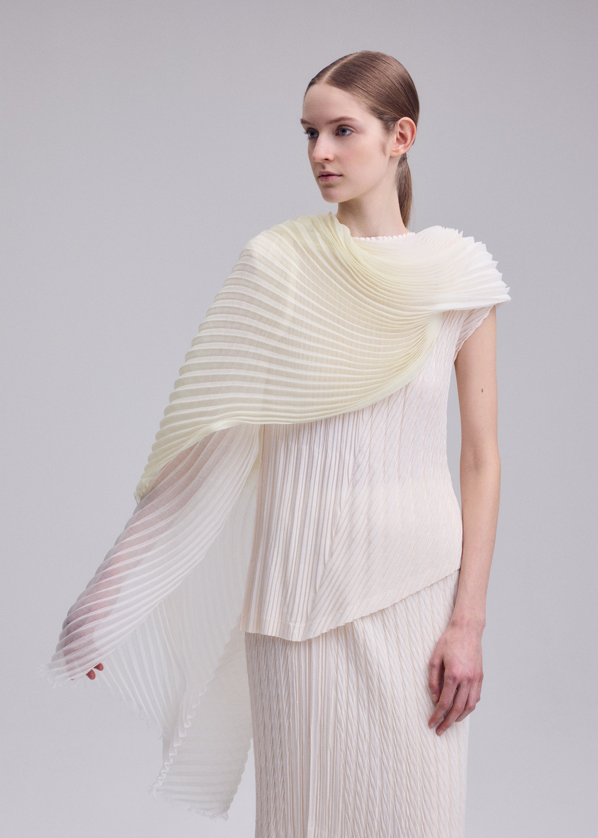 Zoomed view A woman stands against a plain background wearing ISSEY MIYAKE’s GRADATION RAMIE PLEATS STOLE, an asymmetrical, sheer pleated accessory draped over one shoulder, paired with a cream pleated dress. She gazes to the side with her straight brown hair pulled back.