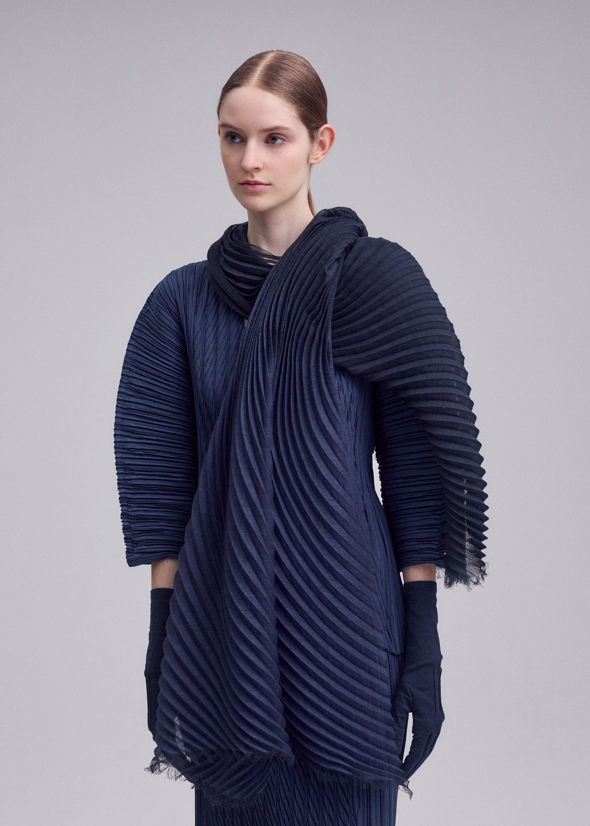 A person with straight, pulled-back hair wears the ISSEY MIYAKE GRADATION RAMIE PLEATS STOLE over a dark, pleated long-sleeve outfit with matching gloves, standing against a plain light background. view 6 of 7