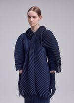 A person with straight, pulled-back hair wears the ISSEY MIYAKE GRADATION RAMIE PLEATS STOLE over a dark, pleated long-sleeve outfit with matching gloves, standing against a plain light background.