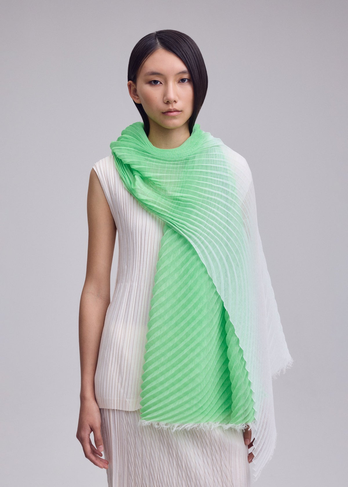 A person with straight black hair in a sleeveless white pleated dress stands against a plain background, draped in the ISSEY MIYAKE GRADATION RAMIE PLEATS STOLE featuring green and white gradation. view 7 of 7
