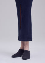 Person wearing a long, dark blue ISSEY MIYAKE ANNA skirt with a front slit and black slip-on shoes, standing on a light-colored surface against a plain background.