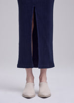 A person wearing the ISSEY MIYAKE ANNA navy blue skirt with a front slit and cream sock-like shoes stands on a light grey background, with only the legs and feet visible.
