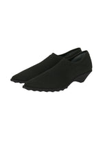 A pair of black, slip-on, pointy-toe shoes with a low wedge heel and textured sole, designed in the signature ISSEY MIYAKE ANNA style, placed on a white background.