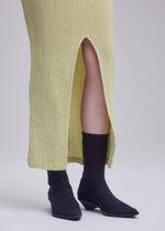 A person wearing a yellow textured skirt with a front slit, black socks, and black ISSEY MIYAKE ANNA shoes, standing on a light-colored surface.