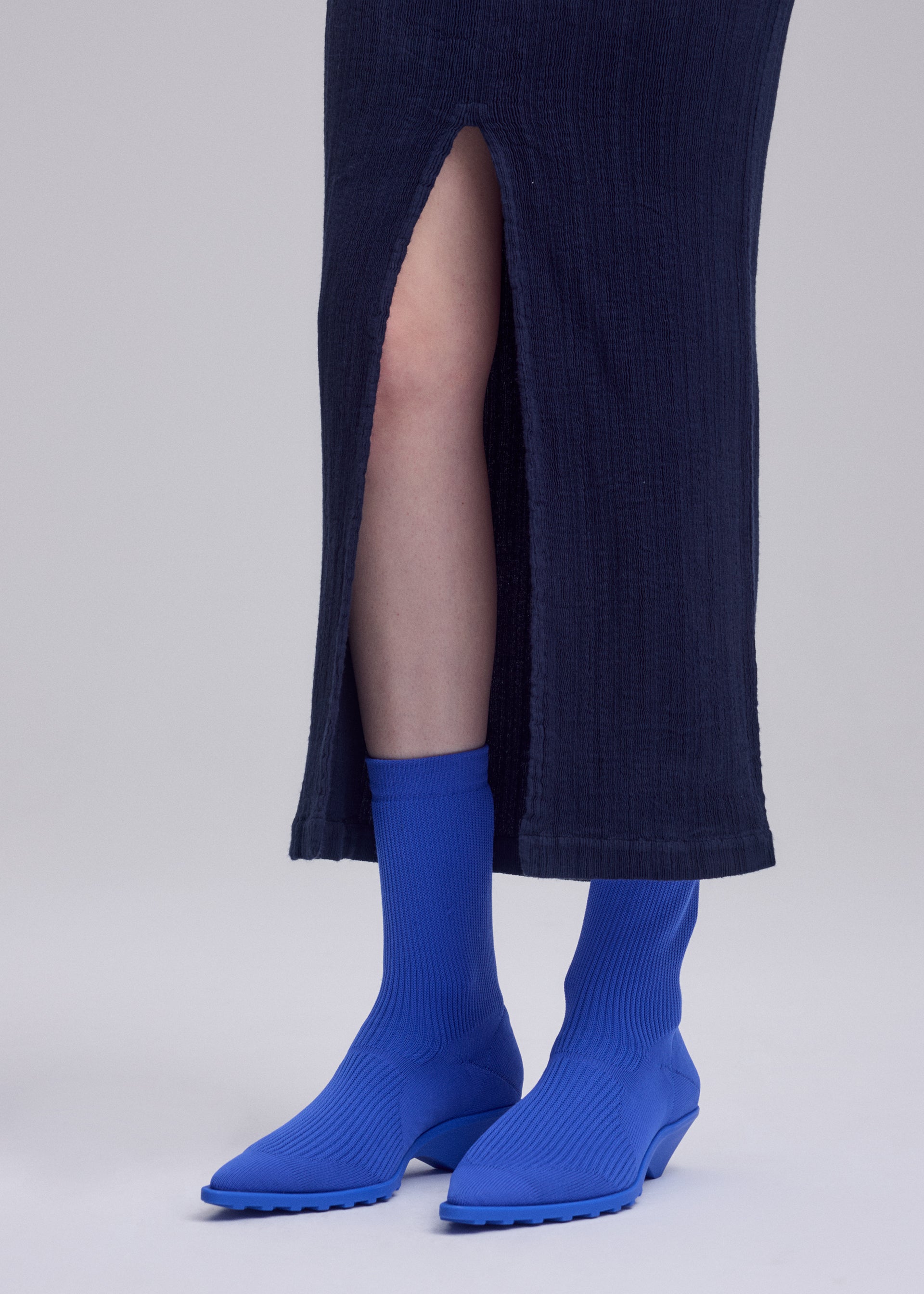 Zoomed view A person wearing a long navy skirt with a front slit, bright blue socks, and matching blue ISSEY MIYAKE ANNA shoes stands on a light gray background.