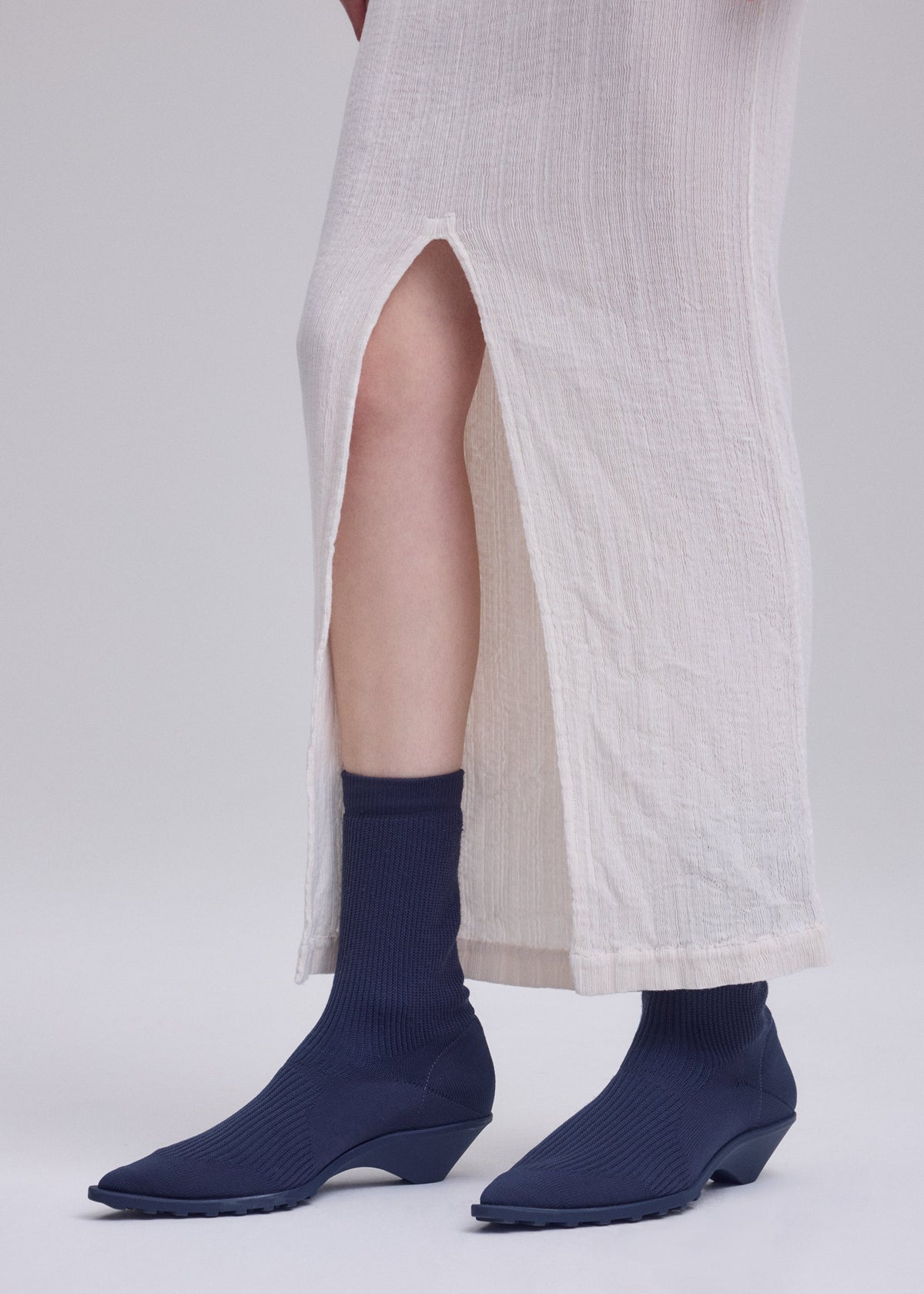 A person wearing a long, off-white textured skirt with a high slit, navy blue ribbed socks, and matching navy blue ISSEY MIYAKE ANNA shoes stands against a plain light background. view 11 of 11