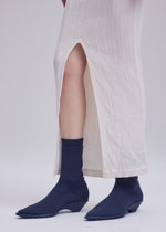 A person wearing a long, off-white textured skirt with a high slit, navy blue ribbed socks, and matching navy blue ISSEY MIYAKE ANNA shoes stands against a plain light background.