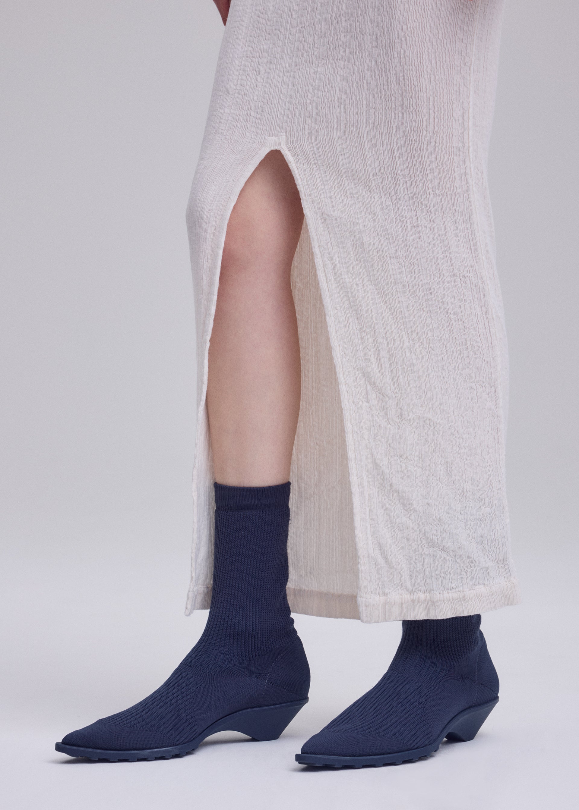 Zoomed view A person wearing a long, off-white textured skirt with a high slit, navy blue ribbed socks, and matching navy blue ISSEY MIYAKE ANNA shoes stands against a plain light background.