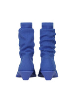 A pair of ISSEY MIYAKE ANNA blue ribbed sock-like boots with geometric, angular heels—reminiscent of knitted footwear—shown from the back against a white background.