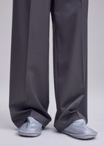 Wearing oversized gray dress pants from ISSEY MIYAKE that drape over most of their feet, the person stands on a gray surface. Only the lower part of the pants and their ISSEY MIYAKE PEU FORM shoes are seen in the image.