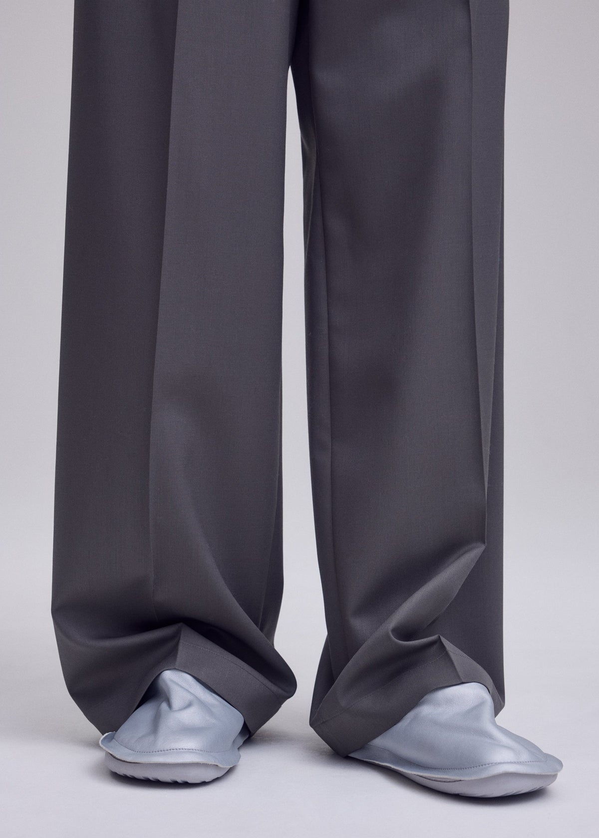 Zoomed view Wearing oversized gray dress pants from ISSEY MIYAKE that drape over most of their feet, the person stands on a gray surface. Only the lower part of the pants and their ISSEY MIYAKE PEU FORM shoes are seen in the image.