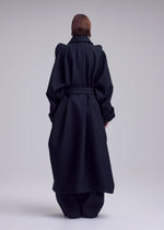 A person with short brown hair is standing with their back to the camera, wearing a long, oversized black CALLIGRAPH wool coat with wide sleeves and a waist belt from ISSEY MIYAKE, in a minimal studio setting.