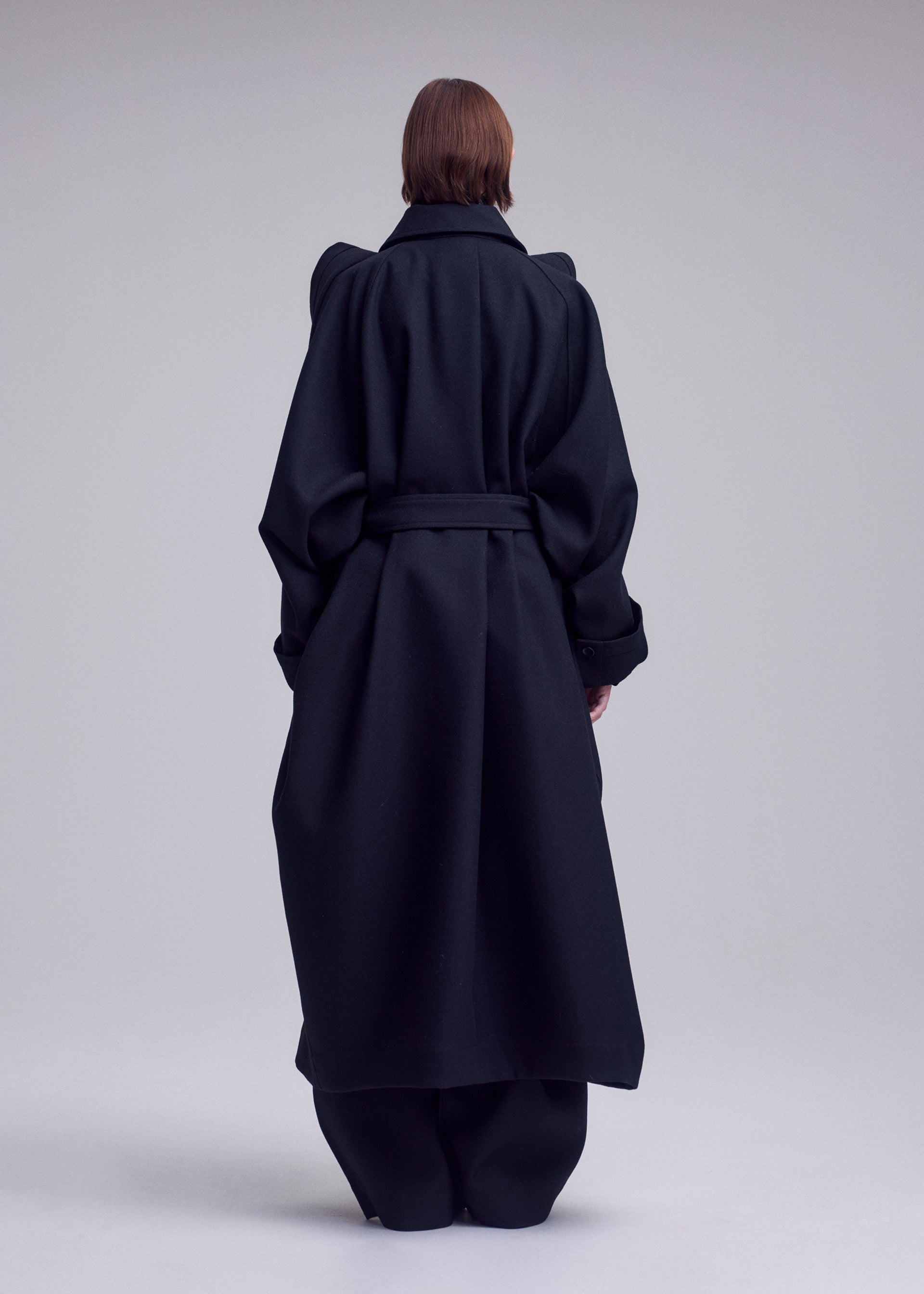 Zoomed view A person with short brown hair is standing with their back to the camera, wearing a long, oversized black CALLIGRAPH wool coat with wide sleeves and a waist belt from ISSEY MIYAKE, in a minimal studio setting.