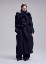 A person stands against a plain background wearing the long, oversized black CALLIGRAPH premium wool coat with a tie belt and wide lapels from ISSEY MIYAKE, paired with black pants and dark shoes. Their hair is straight and tucked behind their ears.