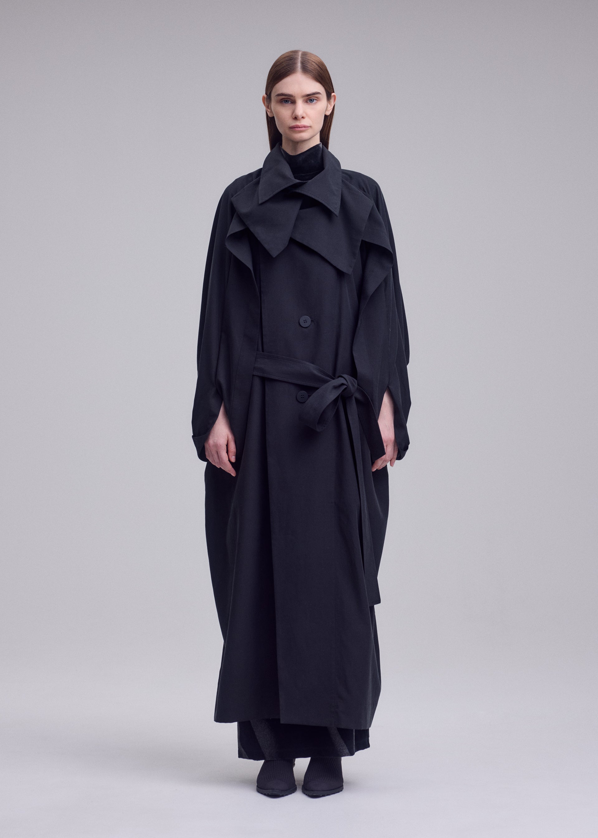 Zoomed view A person stands against a plain background wearing the HANDS OF LOVER long black belted coat with wide lapels, crafted from smooth poplin fabric by ISSEY MIYAKE, paired with matching black shoes. Their hair is straight and tucked behind their ears.