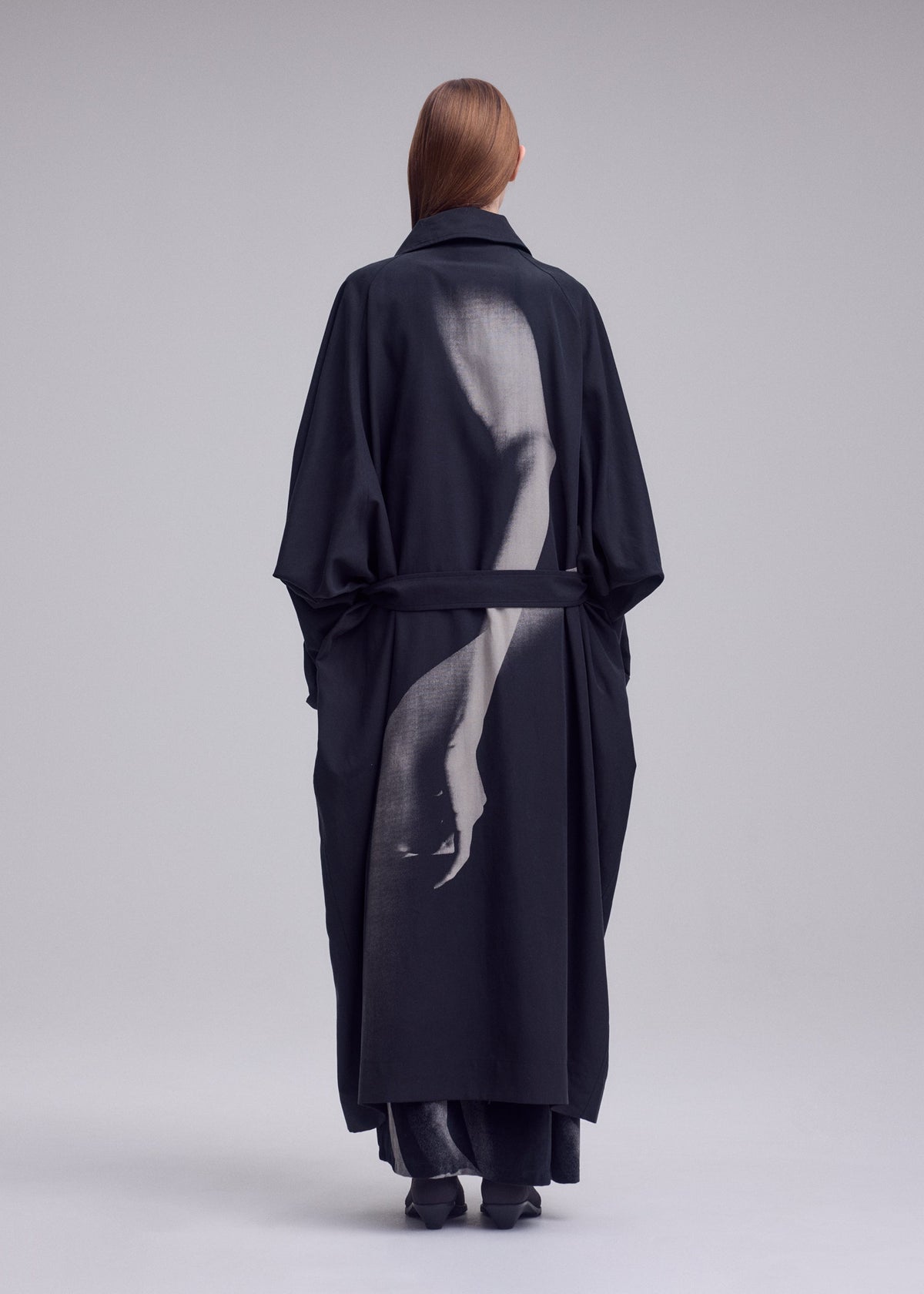A person wearing the ISSEY MIYAKE “HANDS OF LOVER” coat—crafted from smooth poplin fabric and featuring a grayscale silk-screen discharge print of an arm and hand on the back—stands facing away against a neutral background. view 4 of 6
