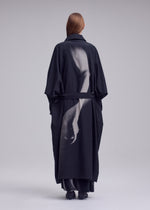 A person wearing the ISSEY MIYAKE “HANDS OF LOVER” coat—crafted from smooth poplin fabric and featuring a grayscale silk-screen discharge print of an arm and hand on the back—stands facing away against a neutral background.